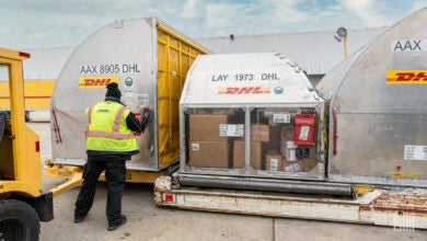 A DHL worker with a yellow vest arranges air containers on the tarmac.