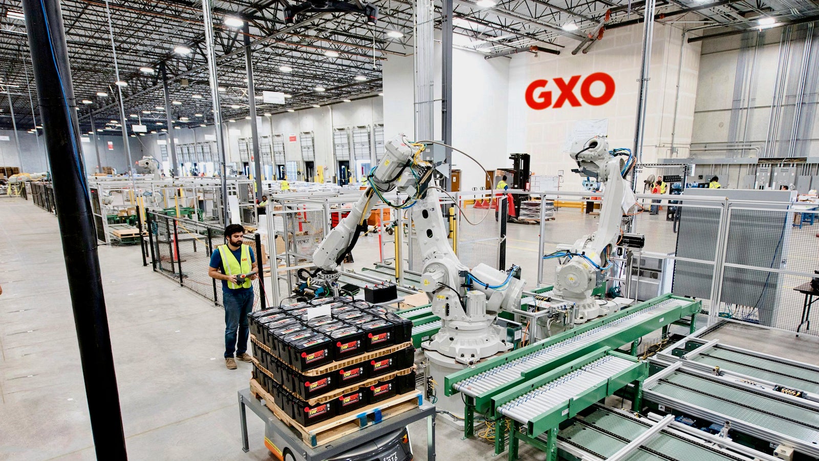 A man works in a warehouse alongside robots while the logo of the company GXO Logistics is visible