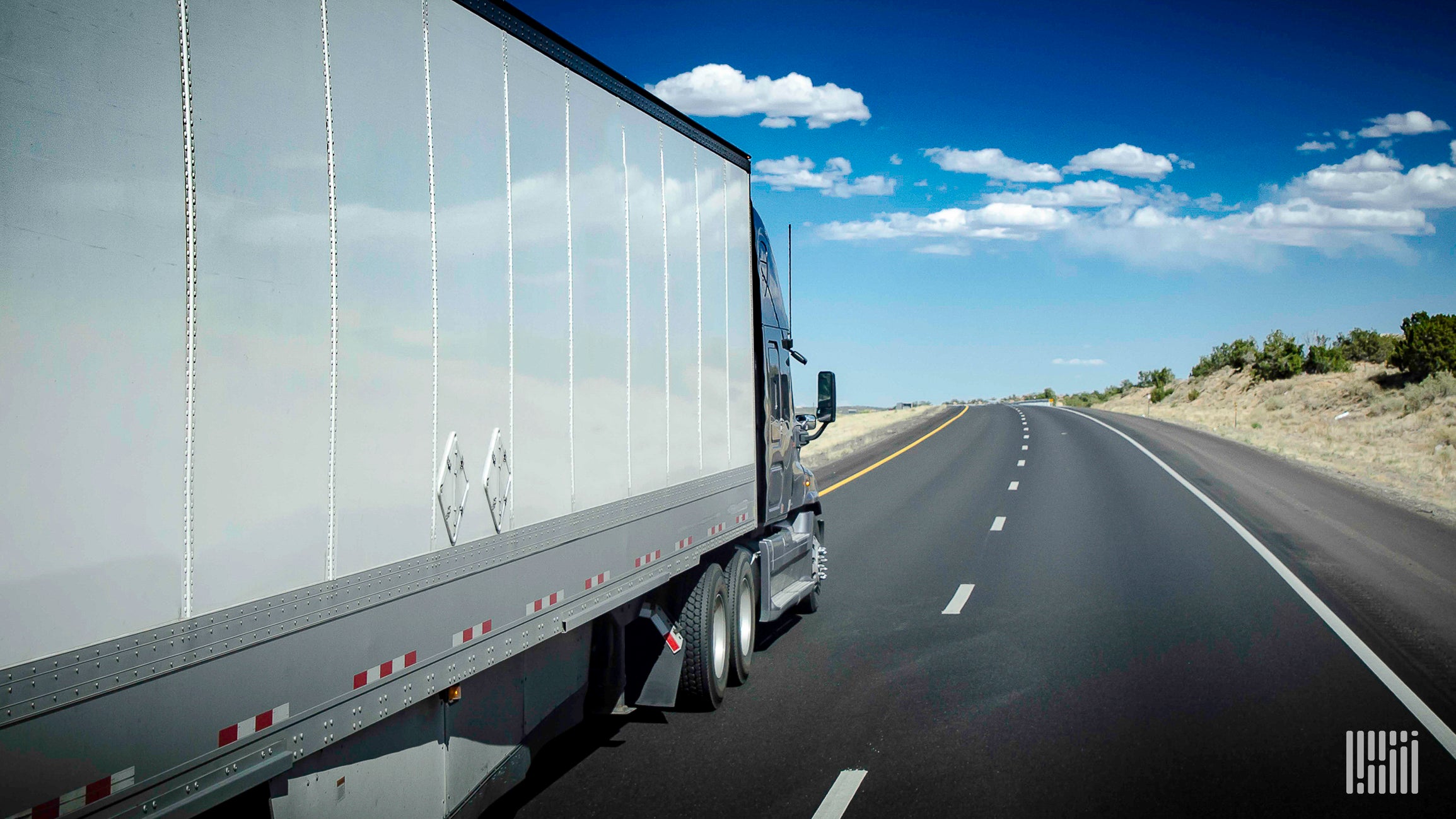 A white semi-truck is traveling down a road with a blue sky in the background.