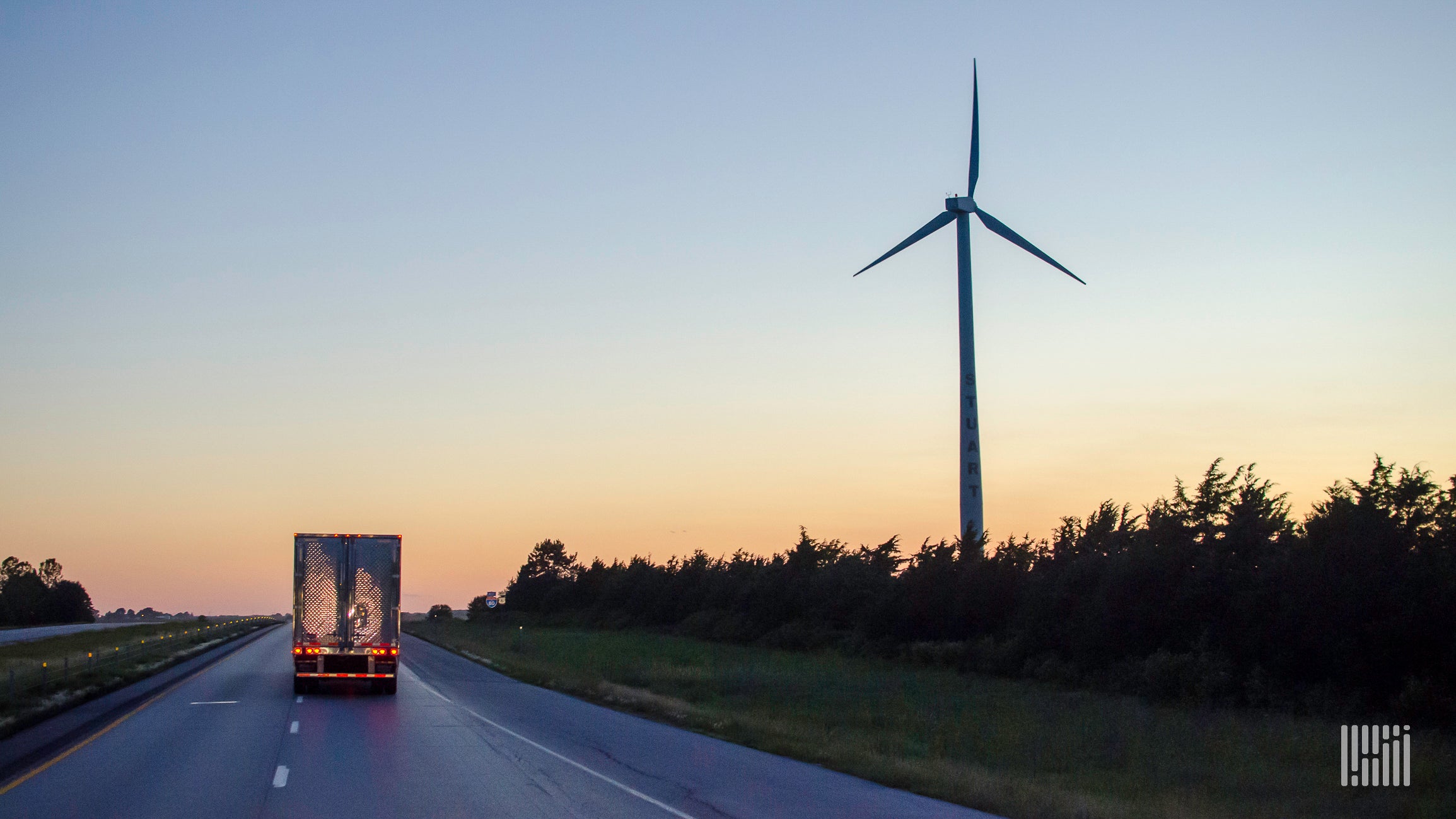 A semi-truck drives down the road with a sunset and wind turbine in the landscape.