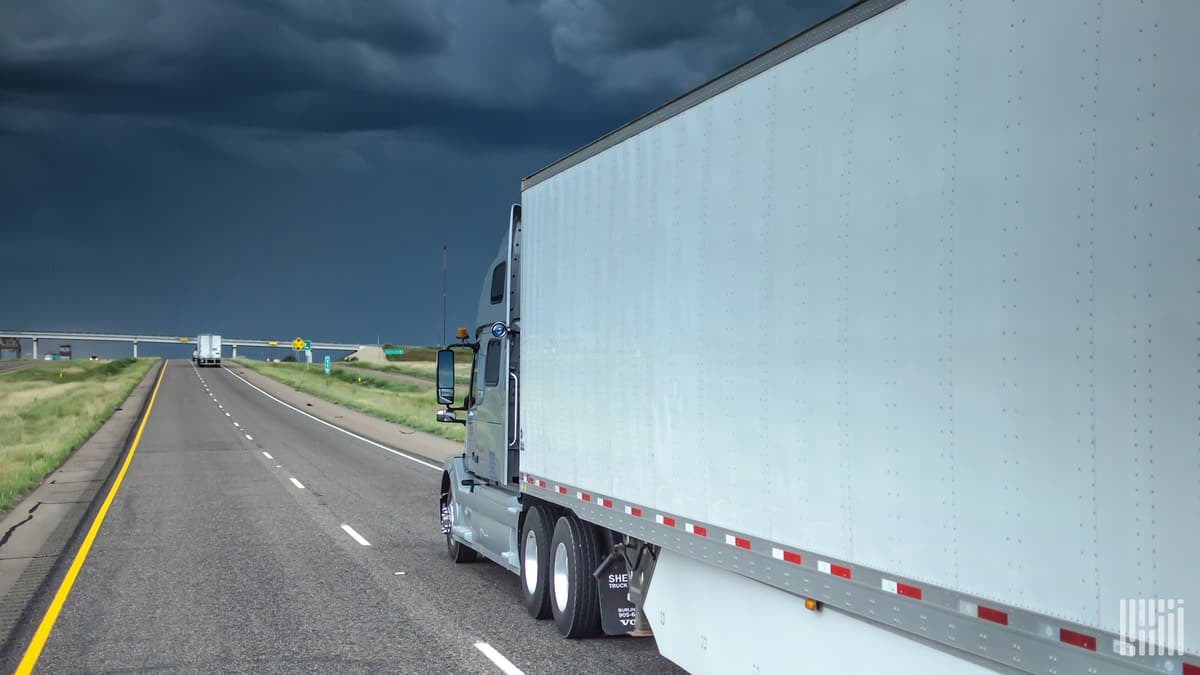 Tractor-trailer heading down a highway with dark storm cloud ahead.