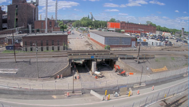 Image shows rail bridge construction, railroad tracks, buildings, sky.